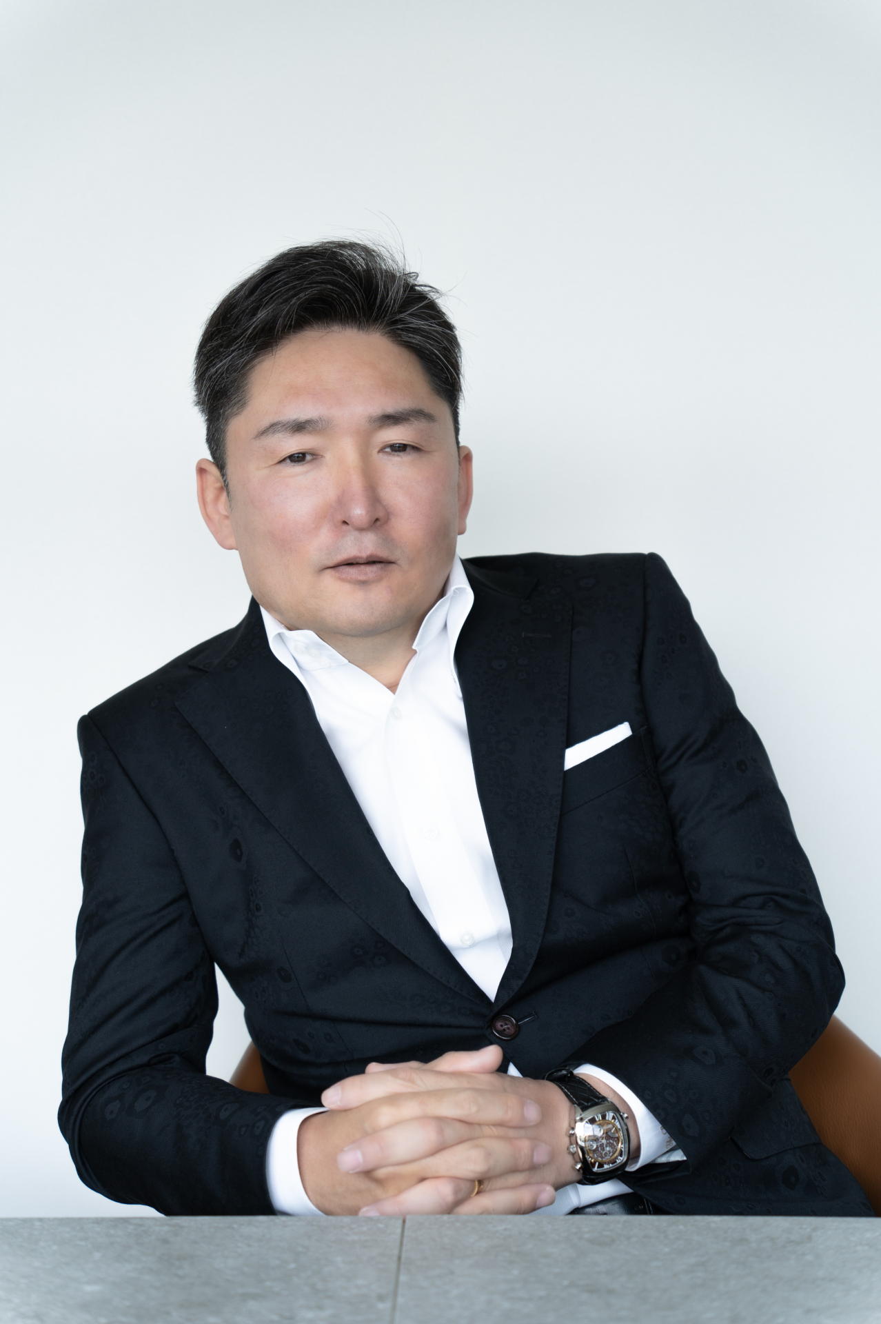 Professional man in a dark suit and white shirt, seated at a table with hands folded, looking at the camera against a plain white background.